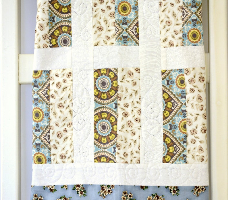 May include: A patchwork quilt featuring vertical strips of fabric with floral, geometric, and circular patterns in shades of blue, yellow, brown, and cream. The design includes white fabric sections with intricate, swirling machine-stitched quilting.
