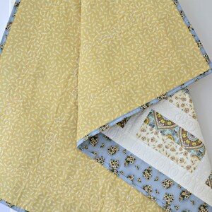 May include: A quilted blanket featuring a mustard yellow fabric with a small white leaf pattern on one side. The reverse side displays a patchwork design with light blue floral fabric, cream-colored sections with intricate stitching, and a border.