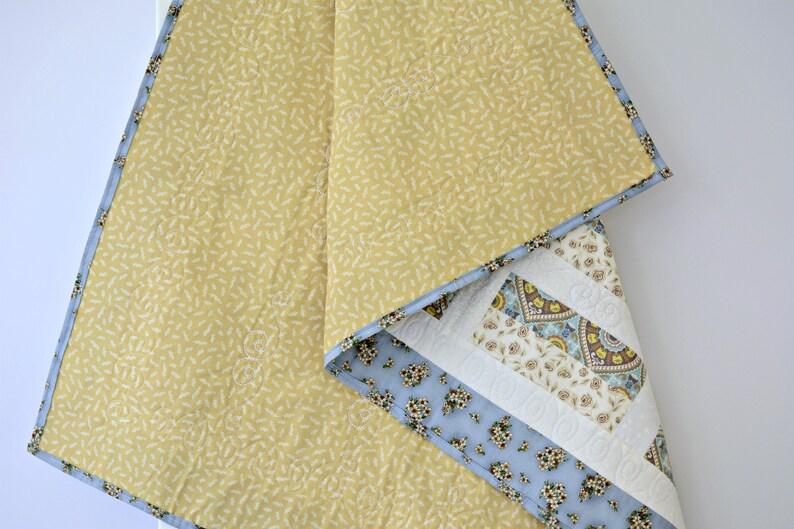 May include: A quilted blanket featuring a mustard yellow fabric with a small white leaf pattern on one side. The reverse side displays a patchwork design with light blue floral fabric, cream-colored sections with intricate stitching, and a border.