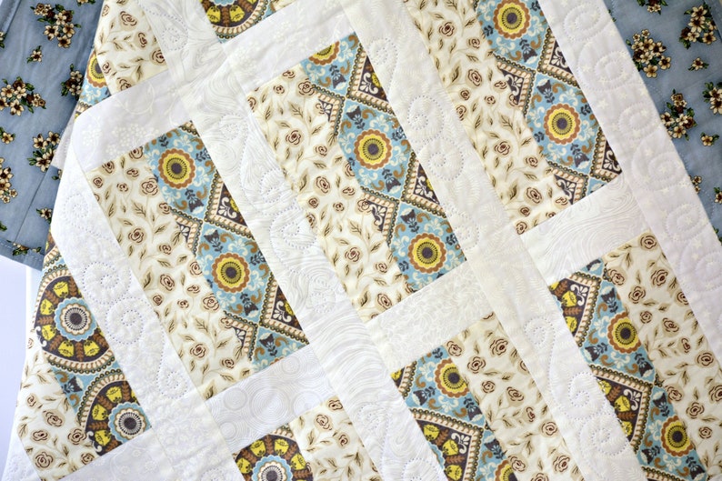 May include: A quilted fabric runner featuring a patchwork design of cream, light blue, and yellow floral and geometric patterns. The textile includes white fabric strips with intricate, swirling machine-stitched quilting details throughout.