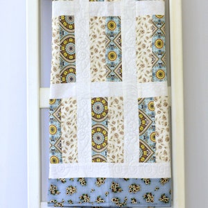 May include: A rectangular quilt featuring vertical panels of cream fabric with brown and blue floral patterns, alternating with white fabric strips displaying intricate quilted swirl designs, finished with a light blue floral border at the bottom.