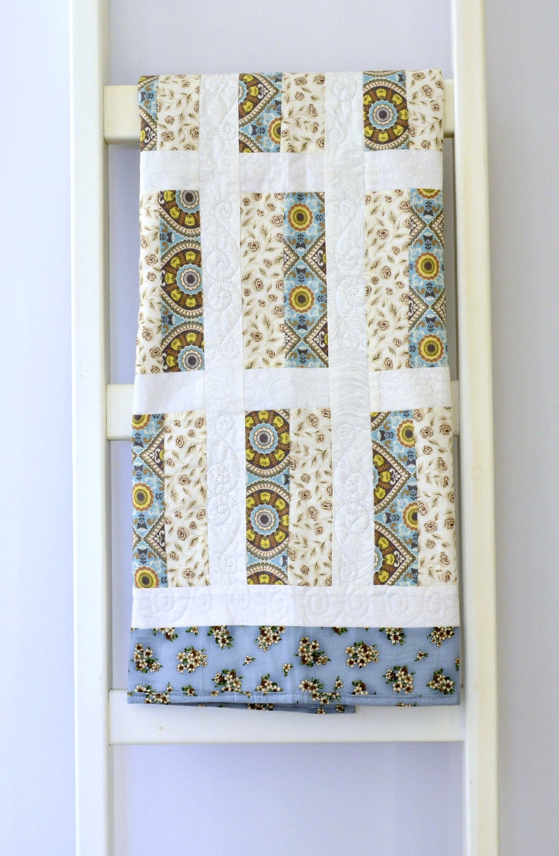 May include: A rectangular quilt featuring vertical panels of cream fabric with brown and blue floral patterns, alternating with white fabric strips displaying intricate quilted swirl designs, finished with a light blue floral border at the bottom.