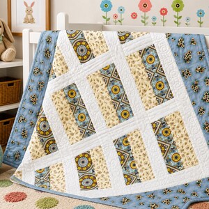May include: A rectangular baby quilt featuring a patchwork design of alternating vertical panels. The quilt displays a repeating pattern of sunflowers, geometric shapes, and floral motifs in shades of blue, yellow, cream, and brown, separated by white fabric strips.