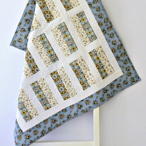 May include: A quilt featuring a central panel of rectangular blocks with cream, blue, and yellow mandala-style patterns separated by white sashing. The quilt is bordered by a light blue fabric with a small, repeating dark floral print pattern.