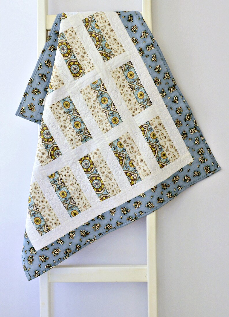 May include: A quilt featuring a central panel of rectangular blocks with cream, blue, and yellow mandala-style patterns separated by white sashing. The quilt is bordered by a light blue fabric with a small, repeating dark floral print pattern.