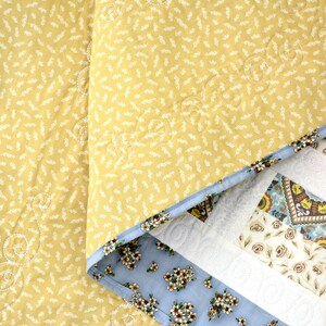 May include: A quilted blanket featuring a mustard yellow top layer with a small white leaf pattern and swirl stitching. The underside reveals a light blue fabric with a floral print and a border of geometric patterns in brown, blue, and white.
