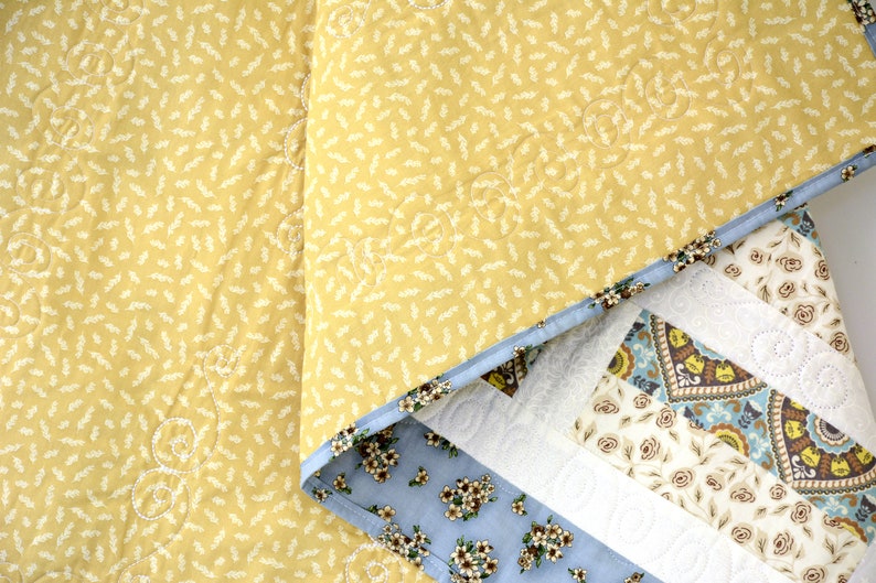 May include: A quilted blanket featuring a mustard yellow top layer with a small white leaf pattern and swirl stitching. The underside reveals a light blue fabric with a floral print and a border of geometric patterns in brown, blue, and white.