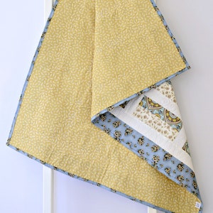 May include: A quilted blanket featuring a mustard yellow top layer with a small white leaf pattern and a light blue binding. The underside reveals a patchwork design with floral patterns in blue, cream, and brown, accented by white quilted swirls.