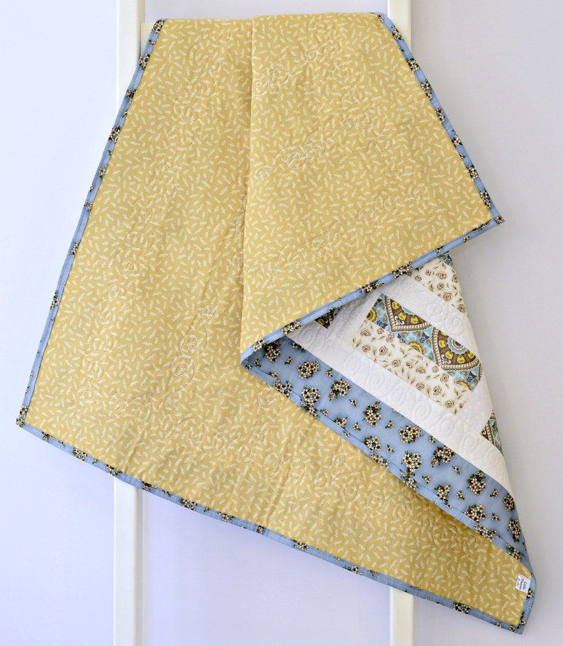 May include: A quilted blanket featuring a mustard yellow top layer with a small white leaf pattern and a light blue binding. The underside reveals a patchwork design with floral patterns in blue, cream, and brown, accented by white quilted swirls.
