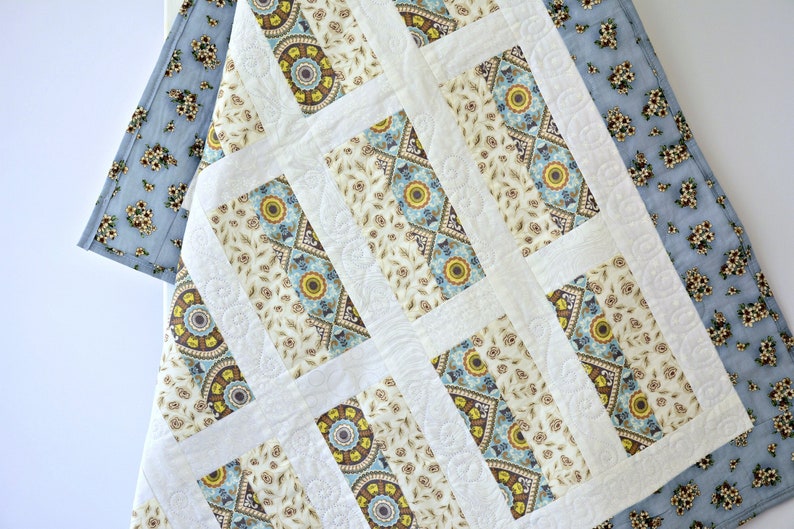May include: A quilted blanket featuring a repeating pattern of rectangular blocks with cream, blue, and yellow floral and geometric motifs. The quilt is bordered by a light blue fabric with a small dark floral print and has white stitched accents.