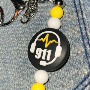 May include: A black and white keychain with a 911 graphic. The keychain has a silver ring and a beaded accent with yellow and white beads. The 911 graphic features a yellow heartbeat line and a headset.
