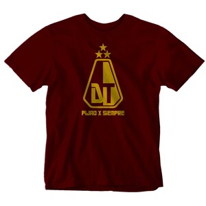 May include: A maroon t-shirt featuring a gold graphic of a shield with the letters "DT" inside. The shield is topped with three gold stars. The text "FIJAD X SIEMPRE" is printed below the shield.