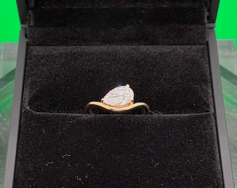 May include: A gold ring with a pear-shaped diamond, presented in a black jewellery box. The ring is displayed on a clear, geometric stand against a green backdrop. The ring's band is a warm gold colour.