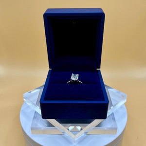 Può includere: Un anello d'argento con un grande diamante a cuscino trasparente incastonato in una fascia semplice. L'anello è esposto in una scatola per anelli in velluto blu.