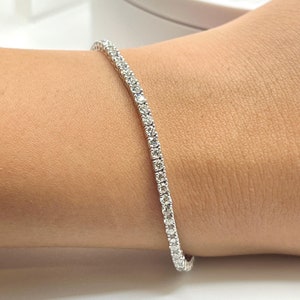 Puede incluir: Un brazalete de tenis delicado con una fila de pequeños diamantes brillantes engastados en una banda de metal blanco.