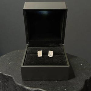 May include: A pair of emerald-cut diamond stud earrings displayed inside a black jewelry box. The earrings are set in a silver or white gold setting. The box is open, revealing the earrings against a black velvet interior.