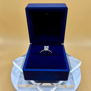 May include: A square-shaped diamond engagement ring with a gold band, sitting inside a blue velvet ring box.