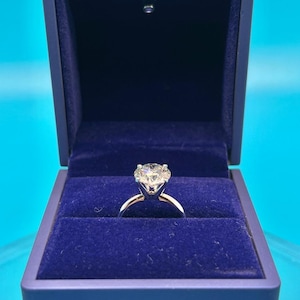 May include: A diamond engagement ring in a blue velvet ring box. The ring has a large, round, brilliant-cut diamond set in a white gold band.