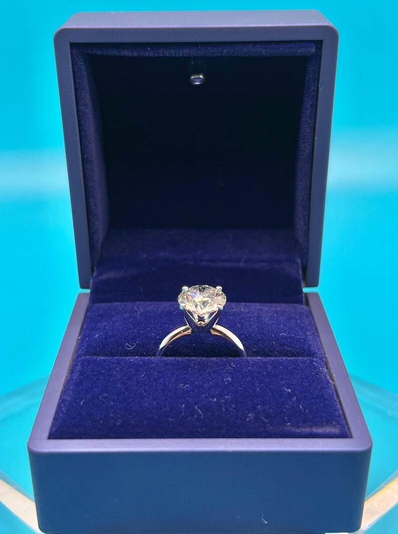 May include: A diamond engagement ring in a blue velvet ring box. The ring has a large, round, brilliant-cut diamond set in a white gold band.