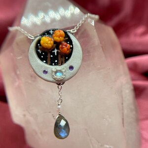 May include: A silver necklace with a crescent moon pendant featuring three orange and yellow berries, a white moonstone, and two purple stones. The pendant is hanging from a silver chain and has a silver drop with a blue labradorite stone.