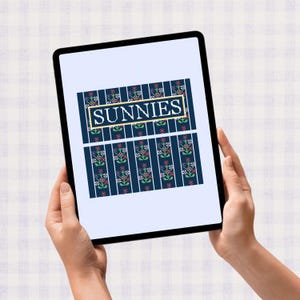 May include: A tablet displays a cross-stitch design with the word "SUNNIES" in a gold frame. The design features a navy blue background with vertical white stripes and floral patterns in red, green, and white.
