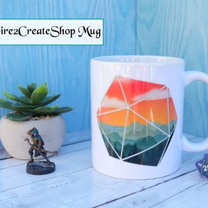 Pode incluir: Caneca de cerâmica branca com um design de paisagem montanhosa colorida dentro de uma forma geométrica que se assemelha a um dado de vinte faces. A caneca está sobre uma superfície de madeira com uma pequena figura e dados.