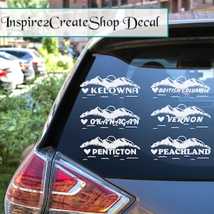 Pode incluir: Autocolantes de vinil branco com desenhos de montanhas e nomes de cidades: Kelowna, British Columbia, Okanagan, Vernon, Penticton e Peachland. O texto "Inspire2CreateShop Decal" está no topo.