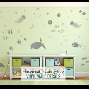 Pode incluir: Um conjunto de adesivos de parede de vinil cinza que representam uma cena subaquática com tartarugas marinhas, águas-vivas, estrelas do mar e peixes. Os adesivos são exibidos em uma parede verde claro em uma sala com uma estante branca e um skate.