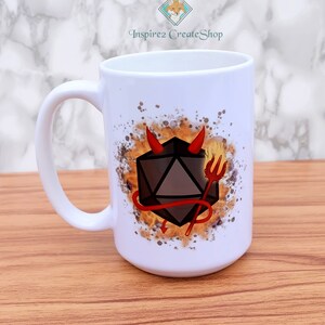 Può includere: Una tazza in ceramica bianca con un'illustrazione nera e rossa di un dado con corna di diavolo e un tridente. Il dado è circondato da un design di fiamme gialle e arancioni.