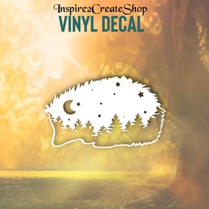 Pode incluir: Autocolante de vinil branco com uma cena de floresta estilizada com árvores, estrelas e uma lua crescente. O autocolante tem o texto "VINYL DECAL" em turquesa. O fundo é uma imagem desfocada de uma floresta.
