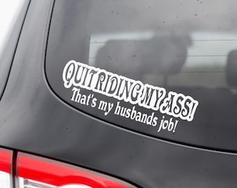 Może przedstawiać: Tylna szyba czarnego samochodu prezentuje białą naklejkę z napisem "QUIT RIDING MY ASS! That's my husbands job!". Światła tylne samochodu są czerwono-białe, na tle czarnego nadwozia.