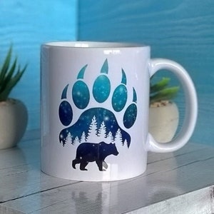 Pode incluir: Caneca de cerâmica branca com um design de pegada de urso azul e preto. A pegada tem um padrão de galáxia com estrelas e uma cena de floresta dentro. Um urso preto está caminhando na cena da floresta. A caneca tem uma alça.