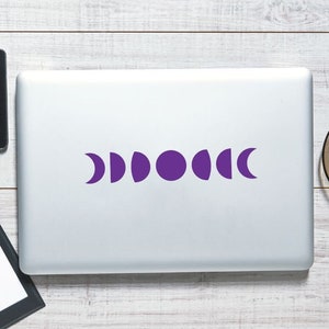 Pode incluir: Um laptop com um design de fases da lua roxo, um smartphone preto, uma prancheta com papel branco em branco, um lápis e uma xícara de café em uma superfície de madeira. O design das fases da lua é uma série de luas crescentes e círculos roxos.