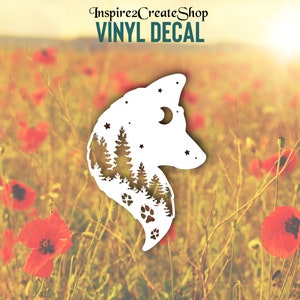 Pode incluir: Autocolante de vinil branco em forma de cabeça de lobo, com uma floresta, estrelas, lua e pegadas. O autocolante está sobre um fundo desfocado de papoilas vermelhas e o texto "Inspire2CreateShop VINYL DECAL".