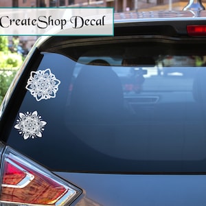 Pode incluir: Duas decalcomanias brancas em estilo mandala estão presas ao vidro traseiro de um carro. As decalcomanias, de tamanhos variados, apresentam padrões simétricos e detalhados. O texto "Inspire2CreateShop Decal" é exibido na parte superior da imagem.
