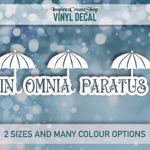 Pode incluir: Autocolante de vinil branco com três desenhos de guarda-chuva acima da frase latina "In Omnia Paratus". O autocolante está sobre um fundo azul e branco desfocado. O texto "2 tamanhos e muitas opções de cores" também está incluído.