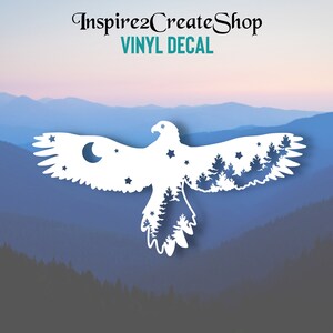 Pode incluir: Vinil branco em forma de águia em voo, com lua crescente, estrelas e árvores incorporadas no design. O texto "Inspire2CreateShop" e "VINYL DECAL" estão no topo. O fundo mostra uma cordilheira.