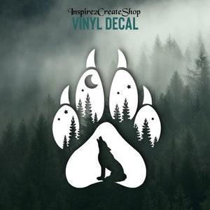 May include: White vinyl decal with a wolf paw print design. The paw print includes a silhouette of a howling wolf, trees, a crescent moon, and stars. The text "Inspire2CreateShop VINYL DECAL" is at the top.