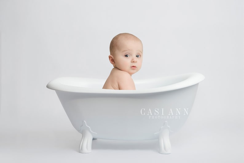 Puede incluir: Un beb&eacute; se sienta en una ba&ntilde;era blanca con un fondo blanco. El beb&eacute; est&aacute; mirando por encima del hombro a la c&aacute;mara.