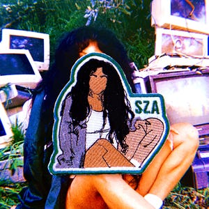 May include: A rectangular embroidered patch featuring a stylized illustration of a person with long dark hair, wearing a light-colored top and a dark jacket. The text SZA is embroidered in green on the right side, bordered by a thick green edge.