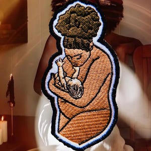 May include: An embroidered patch featuring a brown-toned figure with a high bun hairstyle cradling a small infant. The design is outlined in thick black thread with a thin light blue border, showcasing detailed stitching throughout the figures.