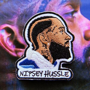 May include: A white and blue embroidered patch with a portrait of a man with a beard and the text "NIPSEY HUSSLE".