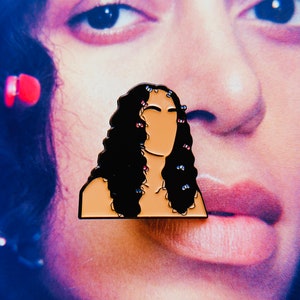 May include: Enamel pin featuring a stylized illustration of a person with long, black curly hair. The pin has a tan face and is adorned with small, colorful hair accessories. The background is a blurred image of a face.