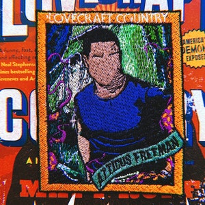 May include: A yellow and orange embroidered patch with a picture of a man in a blue shirt. The text "LOVECRAFT COUNTRY" is at the top of the patch and "ATTICUS FREEMAN" is at the bottom of the patch.