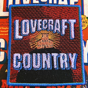 May include: A blue and white embroidered patch with the text "LOVECRAFT COUNTRY" in a stylized font. The patch features a black silhouette of a building with a yellow sunburst behind it.