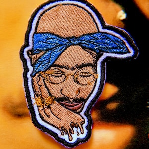 May include: Embroidered patch featuring a portrait of a man with a blue bandana, glasses, and a mustache. The patch has a white and black border. The man's skin is brown, and the bandana is blue.