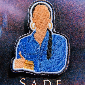 May include: Embroidered patch featuring a portrait of Sade in a blue denim shirt with gold hoop earrings. The patch has a white outline and the word "SADE" is printed below.