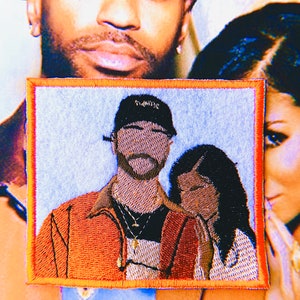 May include: Embroidered patch featuring a couple, the man wearing a black "TWENTYSIX" baseball cap and an orange jacket, the woman with long black hair, against a light blue background.