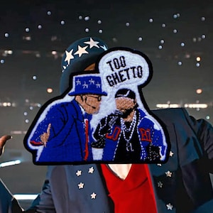 May include: Embroidered patch featuring two figures, one in a blue suit and hat, the other with a microphone. A speech bubble above them reads "TOO GHETTO". The patch has a black border and is set against a blurred background.