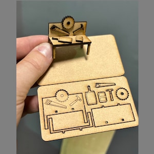 May include: A wooden craft kit featuring a miniature workbench and tools. The kit includes a saw, hammer, screwdriver, and a circular saw blade. The workbench is crafted from wood and has a small shelf. The tools are made of metal and are painted black.